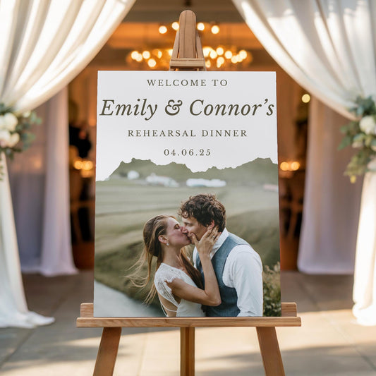 May include: Welcome to Emily & Connor's Rehearsal Dinner 04.06.25 sign with a photo of a couple kissing in front of a mountain range.